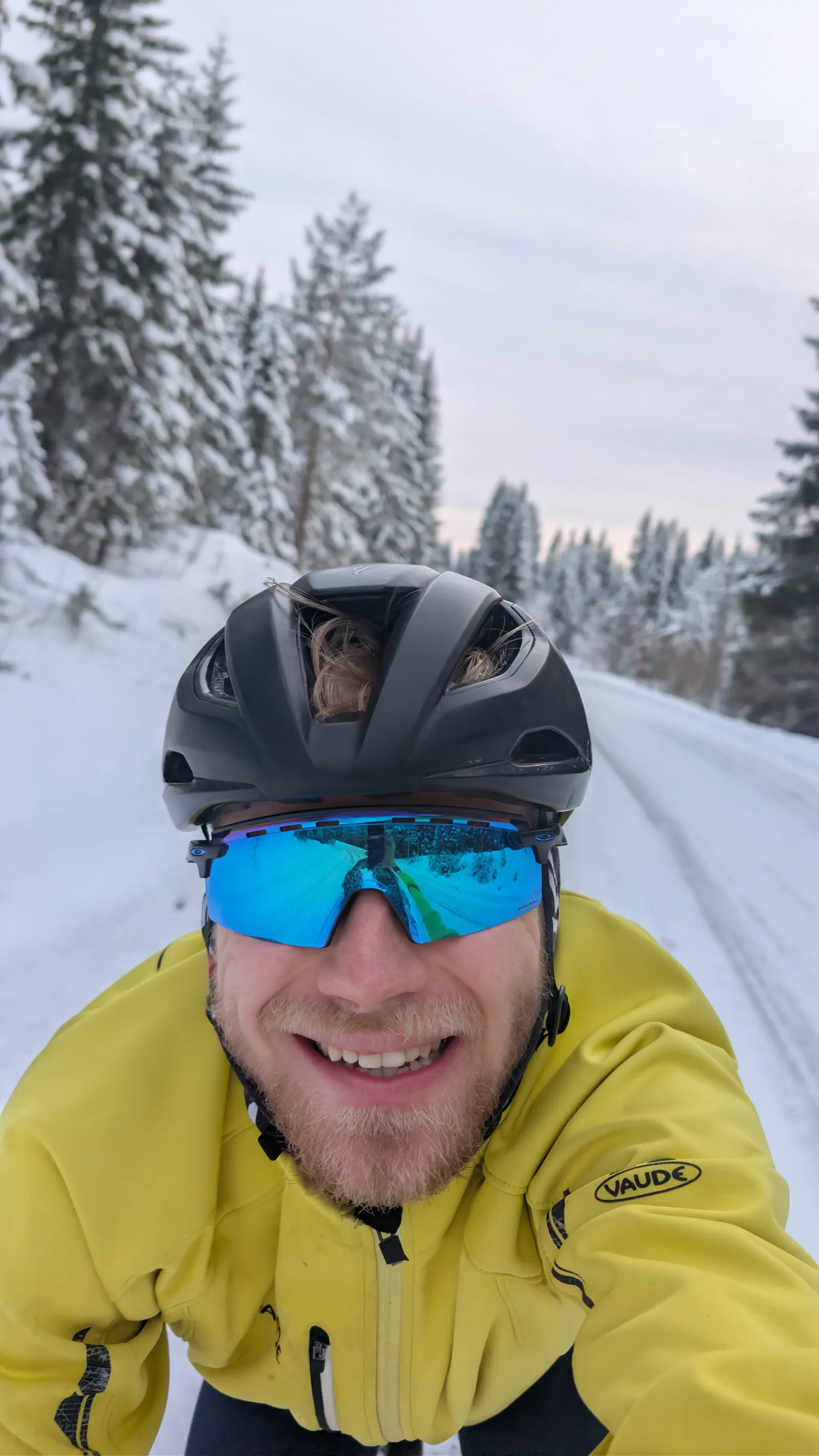 Tycho lachend op de fiets in een geel winterjack, op een besneeuwde weg tussen besneeuwde sparren