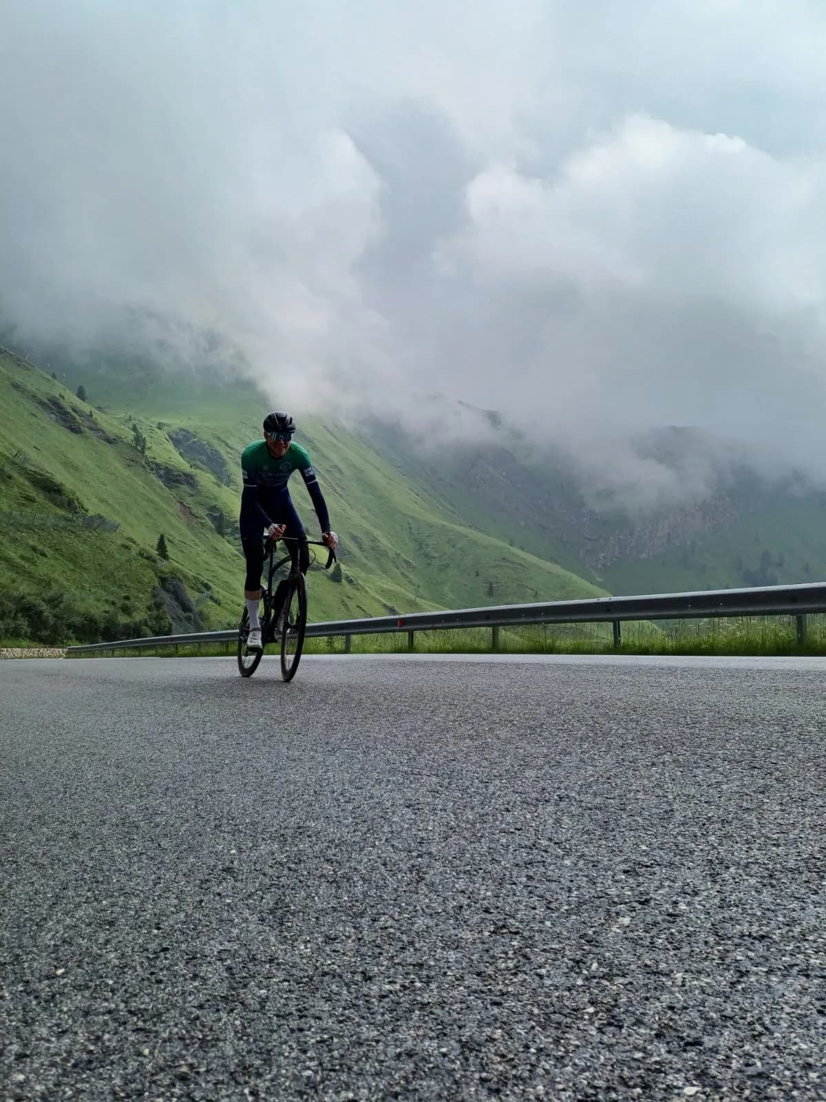 Een renner op een bergpas in Italië, met groene hellingen en laaghangende wolken erachter