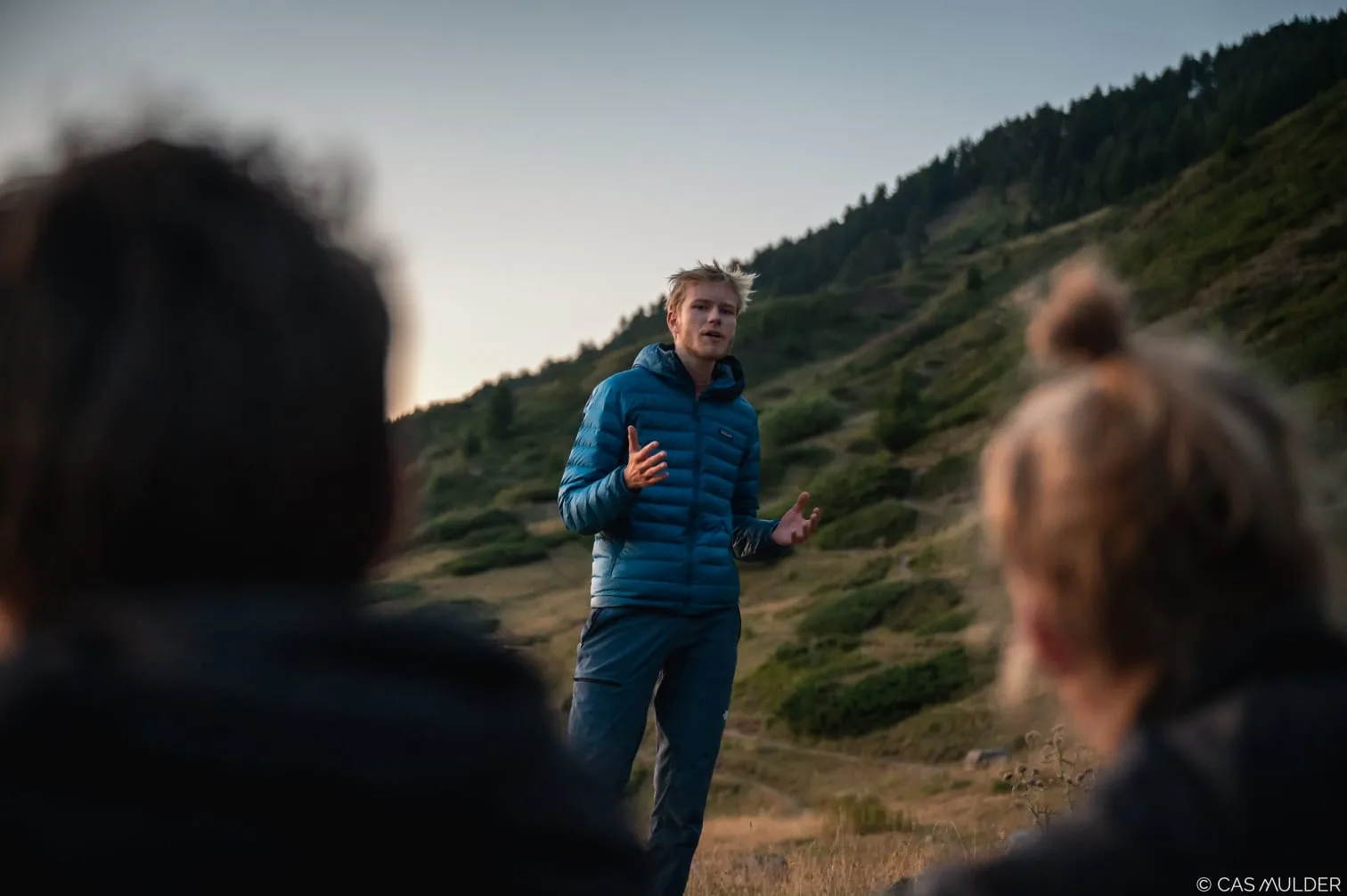 Tycho vertelt een groep over training, buiten op een berghelling bij zonsondergang; twee luisteraars onscherp op de voorgrond